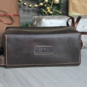 Może przedstawiać: Brązowa sk&oacute;rzana kosmetyczka podr&oacute;żna z zamkiem błyskawicznym i wyszytym monogramem "MDM".