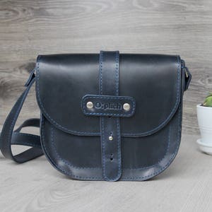 Puede incluir: Bolso bandolera de cuero azul marino con solapa curva y correa ajustable. El bolso presenta costuras azules y cierre de hebilla. La marca "Oplich" est&aacute; grabada en la parte delantera. Una peque&ntilde;a planta en maceta blanca est&aacute; a la derecha.