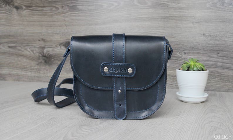 Puede incluir: Bolso bandolera de cuero azul marino con solapa curva y correa ajustable. El bolso presenta costuras azules y cierre de hebilla. La marca "Oplich" est&aacute; grabada en la parte delantera. Una peque&ntilde;a planta en maceta blanca est&aacute; a la derecha.
