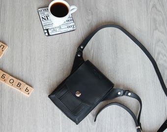 May include: A black leather fanny pack with a silver buckle and a strap. The fanny pack is on a wooden surface with a cup of coffee and a set of wooden tiles spelling out the word "love" in Russian.