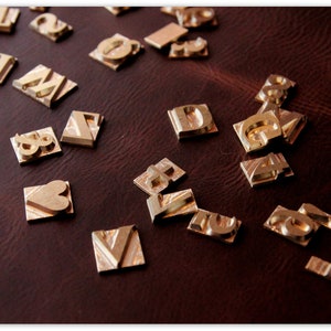 Puede incluir: Una colecci&oacute;n de sellos de letras y n&uacute;meros de metal dorado sobre una superficie de cuero marr&oacute;n. Los sellos est&aacute;n dispuestos en un patr&oacute;n aleatorio e incluyen letras como "A", "D", "G", "M", "V", "X" y n&uacute;meros como "1", "2", "3", "7" y "8". Tambi&eacute;n hay un sello en forma de coraz&oacute;n.