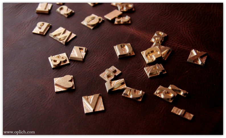 Puede incluir: Una colecci&oacute;n de sellos de letras y n&uacute;meros de metal dorado sobre una superficie de cuero marr&oacute;n. Los sellos est&aacute;n dispuestos en un patr&oacute;n aleatorio e incluyen letras como "A", "D", "G", "M", "V", "X" y n&uacute;meros como "1", "2", "3", "7" y "8". Tambi&eacute;n hay un sello en forma de coraz&oacute;n.