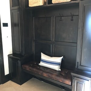 Puede incluir: Un banco de madera oscura con un coj&iacute;n de cuero marr&oacute;n y una almohada a rayas blancas y azules. El banco est&aacute; integrado en un armario de madera oscura con estantes y ganchos.