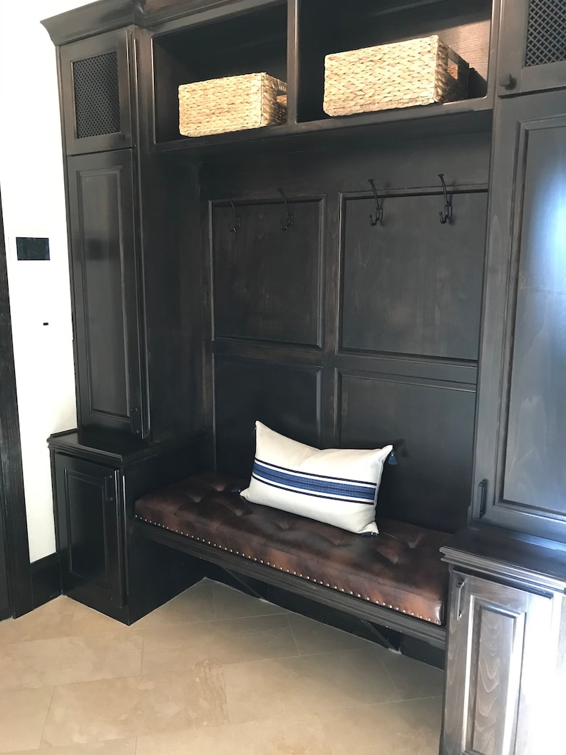 Puede incluir: Un banco de madera oscura con un coj&iacute;n de cuero marr&oacute;n y una almohada a rayas blancas y azules. El banco est&aacute; integrado en un armario de madera oscura con estantes y ganchos.
