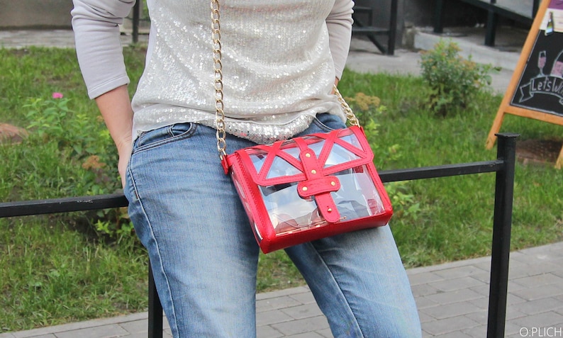 May include: A red and clear plastic crossbody bag with a gold chain strap. The bag has a geometric design and a flap closure with a gold buckle.