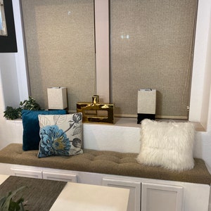 Puede incluir: Un banco de ventana blanco con un coj&iacute;n marr&oacute;n y tres cajones blancos. El banco tiene dos almohadas blancas, una con un patr&oacute;n floral y la otra con una textura blanca esponjosa. El banco est&aacute; frente a una ventana con persianas beige.