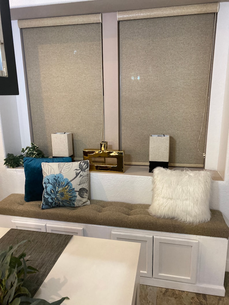 Puede incluir: Un banco de ventana blanco con un coj&iacute;n marr&oacute;n y tres cajones blancos. El banco tiene dos almohadas blancas, una con un patr&oacute;n floral y la otra con una textura blanca esponjosa. El banco est&aacute; frente a una ventana con persianas beige.