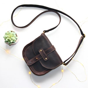 Puede incluir: Bolso bandolera de cuero marr&oacute;n oscuro con solapa curva y correa ajustable. El bolso tiene un cierre de hebilla y costuras decorativas. Se ve una peque&ntilde;a planta suculenta en una maceta blanca.
