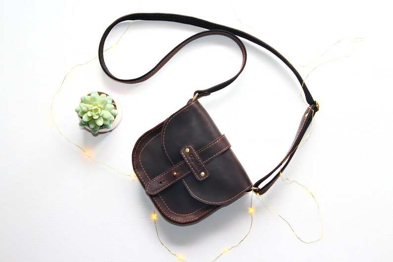 Puede incluir: Bolso bandolera de cuero marr&oacute;n oscuro con solapa curva y correa ajustable. El bolso tiene un cierre de hebilla y costuras decorativas. Se ve una peque&ntilde;a planta suculenta en una maceta blanca.