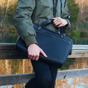 May include: A person carrying a black leather briefcase with a gold zipper and hardware. The briefcase has a strap and is being held by the handle.