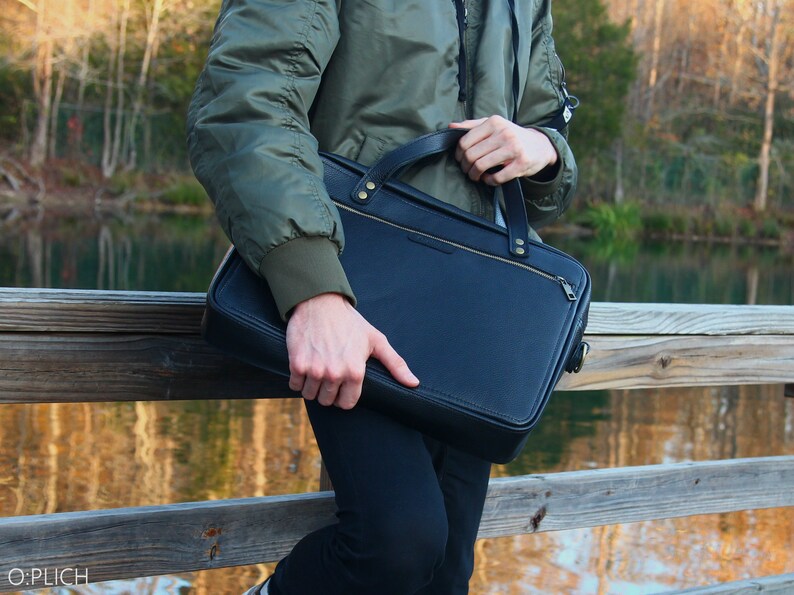 May include: A person carrying a black leather briefcase with a gold zipper and hardware. The briefcase has a strap and is being held by the handle.