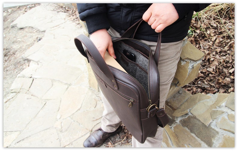 May include: A brown leather briefcase with a zippered closure and a flap. The briefcase has a handle and a shoulder strap. The interior is lined with a gray fabric.