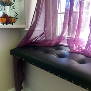 Puede incluir: Un asiento de ventana de cuero negro con tapicer&iacute;a capiton&eacute; y ribete de tachuelas de lat&oacute;n. El asiento est&aacute; colocado debajo de una ventana con cortinas de voile morado.