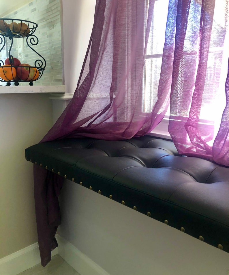 Puede incluir: Un asiento de ventana de cuero negro con tapicer&iacute;a capiton&eacute; y ribete de tachuelas de lat&oacute;n. El asiento est&aacute; colocado debajo de una ventana con cortinas de voile morado.