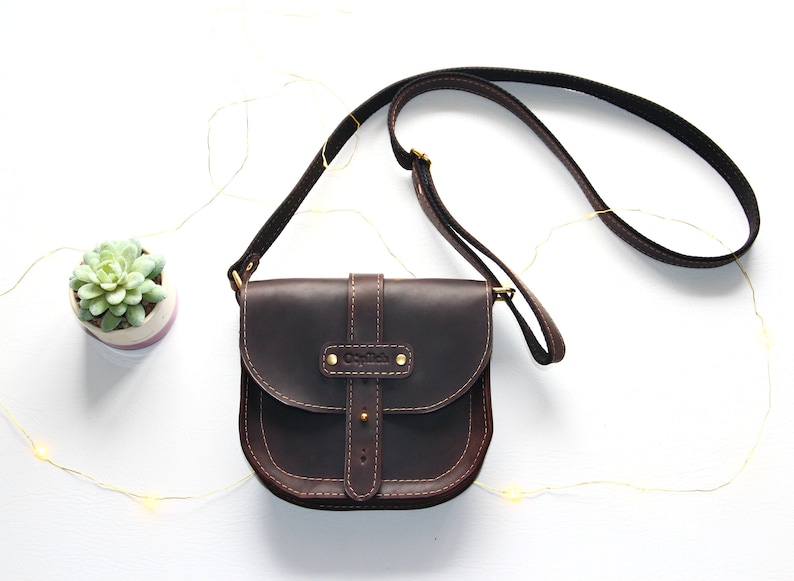Puede incluir: Bolso bandolera de cuero marr&oacute;n oscuro con cierre de solapa y correa ajustable. El bolso presenta costuras visibles y herrajes dorados. El nombre de la marca "Craftish" est&aacute; grabado en una peque&ntilde;a placa rectangular.