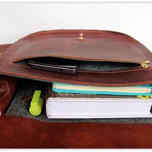 Puede incluir: Bolso mensajero de cuero marr&oacute;n con solapa y bandolera. El bolso est&aacute; abierto, revelando un bolsillo con cremallera, un smartphone, cuadernos y un resaltador. Dise&ntilde;ado para llevar lo esencial del d&iacute;a a d&iacute;a.