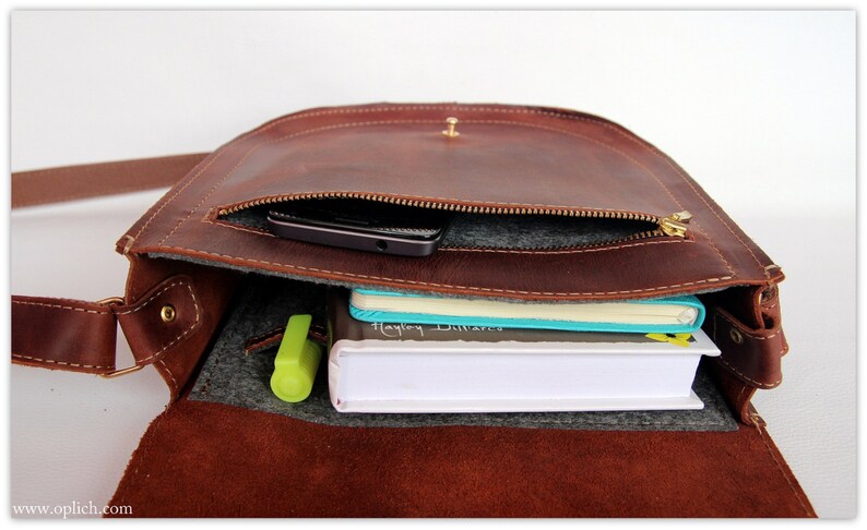 Puede incluir: Bolso mensajero de cuero marr&oacute;n con solapa y bandolera. El bolso est&aacute; abierto, revelando un bolsillo con cremallera, un smartphone, cuadernos y un resaltador. Dise&ntilde;ado para llevar lo esencial del d&iacute;a a d&iacute;a.
