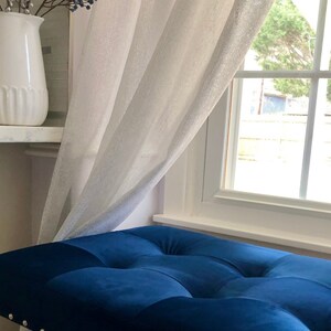 Puede incluir: Un banco tapizado en terciopelo azul con tachuelas plateadas delante de una ventana con cortinas blancas de voile.