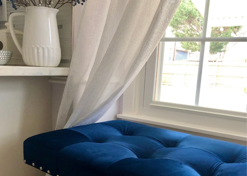 Puede incluir: Un banco tapizado en terciopelo azul con tachuelas plateadas delante de una ventana con cortinas blancas de voile.