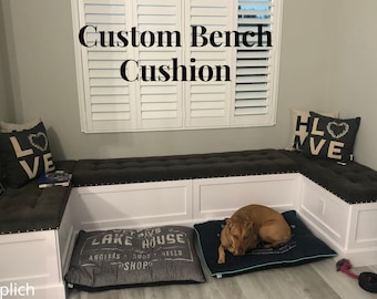 May include: A custom-made bench cushion with a gray fabric cover and tufted buttons. The bench is in a corner and has white trim. There is a dog bed with a gray and white patterned cover in front of the bench. A dog is lying on the bed. The text "Custom Bench Cushion" is written above the bench.