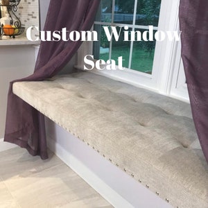 May include: A custom window seat with a light gray upholstered cushion and silver tacks. The seat is positioned under a window with white trim and purple curtains.