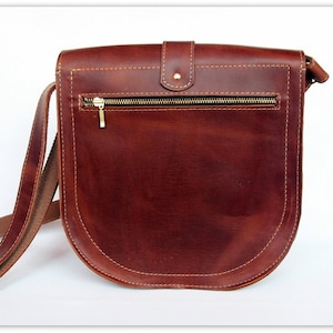 Puede incluir: Bolso bandolera de cuero marr&oacute;n con base curvada y cierre de solapa. El bolso tiene un bolsillo frontal con cremallera dorada y una correa larga y ajustable. Las costuras son de un color m&aacute;s claro.