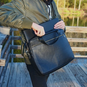 May include: A black leather briefcase with a zipper closure and a gold handle. The briefcase is being held by a person wearing a green jacket.