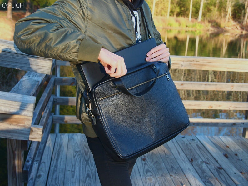 May include: A black leather briefcase with a zipper closure and a gold handle. The briefcase is being held by a person wearing a green jacket.