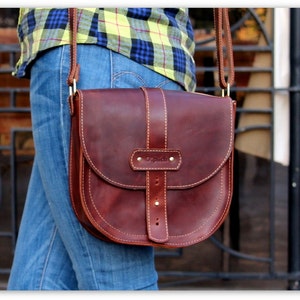 Puede incluir: Bolso bandolera de cuero marr&oacute;n con solapa curva y correa ajustable. El bolso tiene un cierre de hebilla y costuras decorativas. La palabra "Oplich" est&aacute; grabada en la parte delantera. El bolso se lleva al hombro.