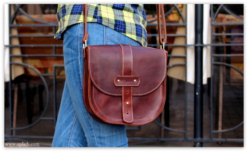 Puede incluir: Bolso bandolera de cuero marr&oacute;n con solapa curva y correa ajustable. El bolso tiene un cierre de hebilla y costuras decorativas. La palabra "Oplich" est&aacute; grabada en la parte delantera. El bolso se lleva al hombro.