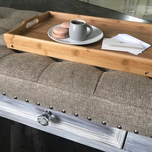 May include: A wooden serving tray with a cup of coffee, two macarons, and a white card on a gray upholstered ottoman.