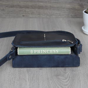 Puede incluir: Un bolso bandolera de cuero azul marino con cremallera y correa plateadas. El bolso est&aacute; abierto, revelando un libro verde con el t&iacute;tulo "PRINCESS" en letras blancas. Una peque&ntilde;a planta suculenta en una maceta blanca est&aacute; en el fondo.