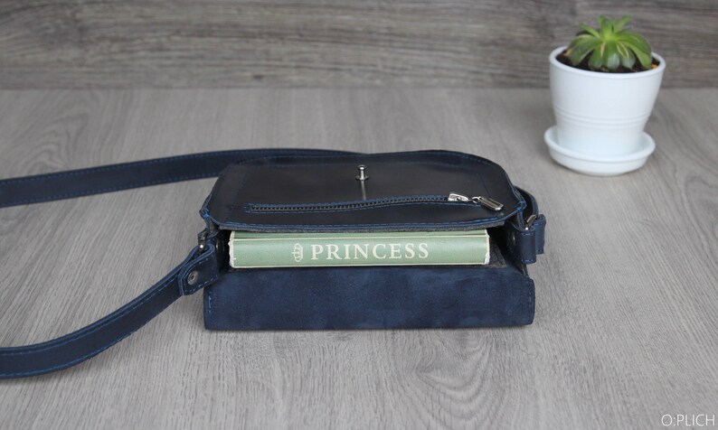 Puede incluir: Un bolso bandolera de cuero azul marino con cremallera y correa plateadas. El bolso est&aacute; abierto, revelando un libro verde con el t&iacute;tulo "PRINCESS" en letras blancas. Una peque&ntilde;a planta suculenta en una maceta blanca est&aacute; en el fondo.