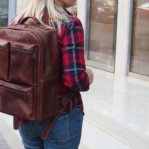 Puede incluir: Una mochila de cuero marr&oacute;n con varios bolsillos y cierre de cremallera. La mochila la lleva puesta una persona que lleva una camisa de cuadros rojos y negros y unos vaqueros azules.