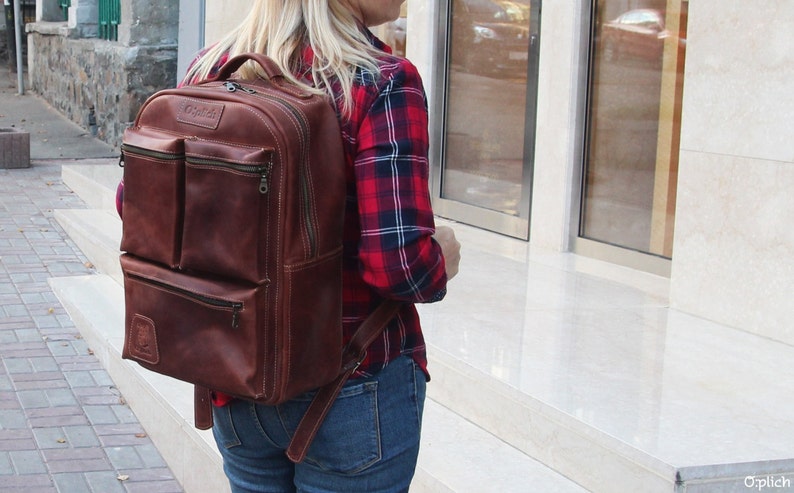 Puede incluir: Una mochila de cuero marr&oacute;n con varios bolsillos y cierre de cremallera. La mochila la lleva puesta una persona que lleva una camisa de cuadros rojos y negros y unos vaqueros azules.