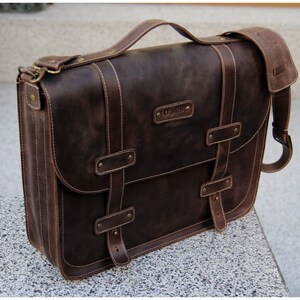 K&ouml;nnte beinhalten: Eine braune Ledertasche mit einem langen verstellbaren Schultergurt, zwei Schnallen und einem Tragegriff. Die Tasche hat eine Klappe mit einem Schnallenverschluss und ein kleines Fach auf der Vorderseite. Die Tasche hat einen gepr&auml;gten Markennamen auf der Vorderseite.