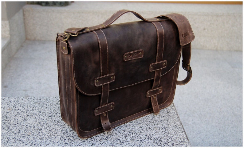 K&ouml;nnte beinhalten: Eine braune Ledertasche mit einem langen verstellbaren Schultergurt, zwei Schnallen und einem Tragegriff. Die Tasche hat eine Klappe mit einem Schnallenverschluss und ein kleines Fach auf der Vorderseite. Die Tasche hat einen gepr&auml;gten Markennamen auf der Vorderseite.