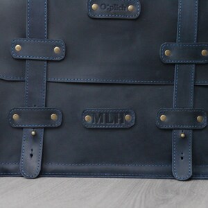 K&ouml;nnte beinhalten: Eine dunkelblaue Ledertasche mit Messingbeschl&auml;gen und blauen N&auml;hten. Die Tasche hat zwei Riemen mit Schnallen und zwei Namensschilder, eines mit der Aufschrift "O'plich" und das andere mit der Aufschrift "MLH".