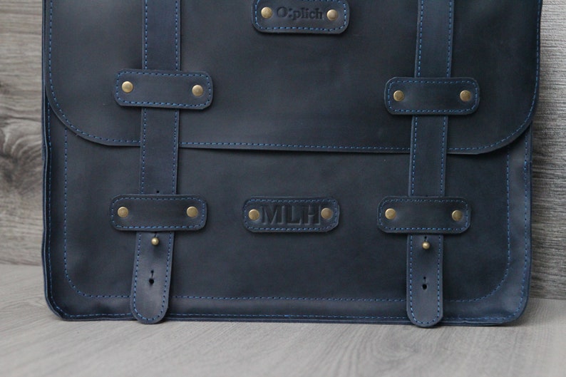 K&ouml;nnte beinhalten: Eine dunkelblaue Ledertasche mit Messingbeschl&auml;gen und blauen N&auml;hten. Die Tasche hat zwei Riemen mit Schnallen und zwei Namensschilder, eines mit der Aufschrift "O'plich" und das andere mit der Aufschrift "MLH".