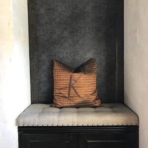 Puede incluir: Un banco tapizado gris con un marco de madera negro y dos cajones. El banco tiene un coj&iacute;n marr&oacute;n con un gran monograma "K" en &eacute;l. La pared detr&aacute;s del banco est&aacute; cubierta con un papel tapiz texturizado gris oscuro.