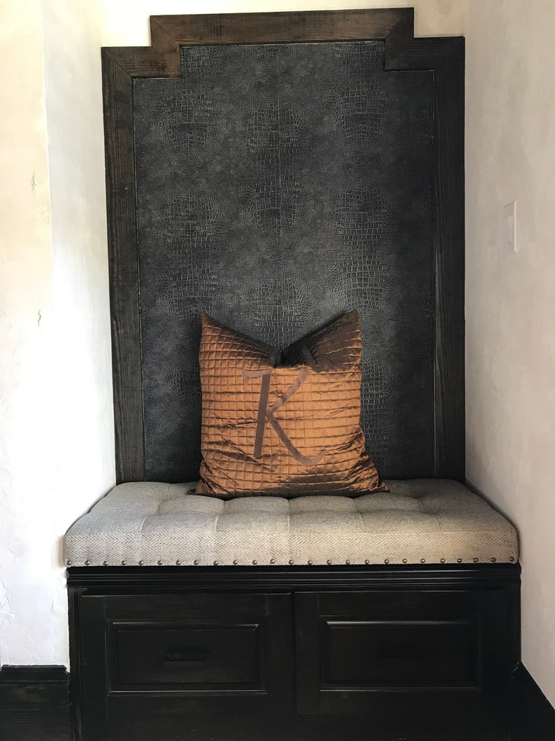 Puede incluir: Un banco tapizado gris con un marco de madera negro y dos cajones. El banco tiene un coj&iacute;n marr&oacute;n con un gran monograma "K" en &eacute;l. La pared detr&aacute;s del banco est&aacute; cubierta con un papel tapiz texturizado gris oscuro.