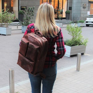 Puede incluir: Una mujer con una camisa de cuadros rojos y negros camina por una calle de la ciudad. Lleva una mochila de cuero marr&oacute;n con varios bolsillos y cremalleras.