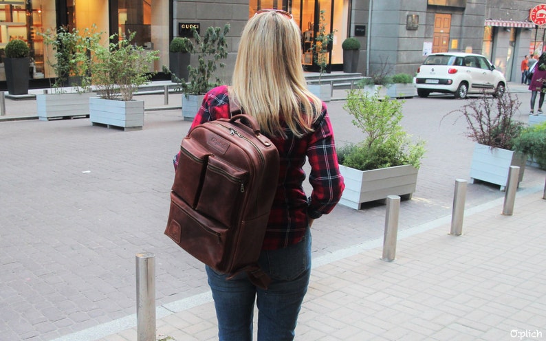 Puede incluir: Una mujer con una camisa de cuadros rojos y negros camina por una calle de la ciudad. Lleva una mochila de cuero marr&oacute;n con varios bolsillos y cremalleras.