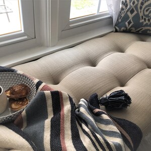 Puede incluir: Un acogedor asiento de ventana con una manta de rayas, un taz&oacute;n de panqueques y una taza de caf&eacute;. El asiento est&aacute; tapizado en una tela beige claro con botones.