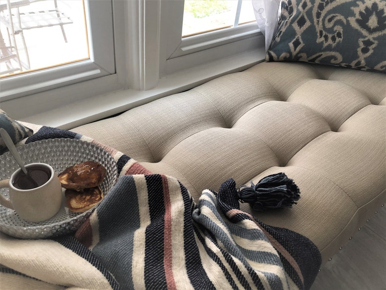 Puede incluir: Un acogedor asiento de ventana con una manta de rayas, un taz&oacute;n de panqueques y una taza de caf&eacute;. El asiento est&aacute; tapizado en una tela beige claro con botones.