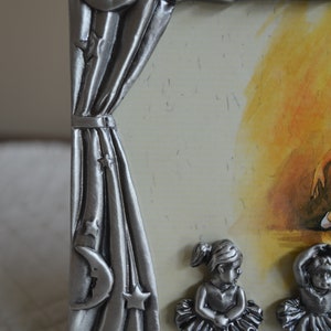 May include: Silver-tone picture frame with a curtain design and stars. The frame features three ballerina figurines in the bottom corners.