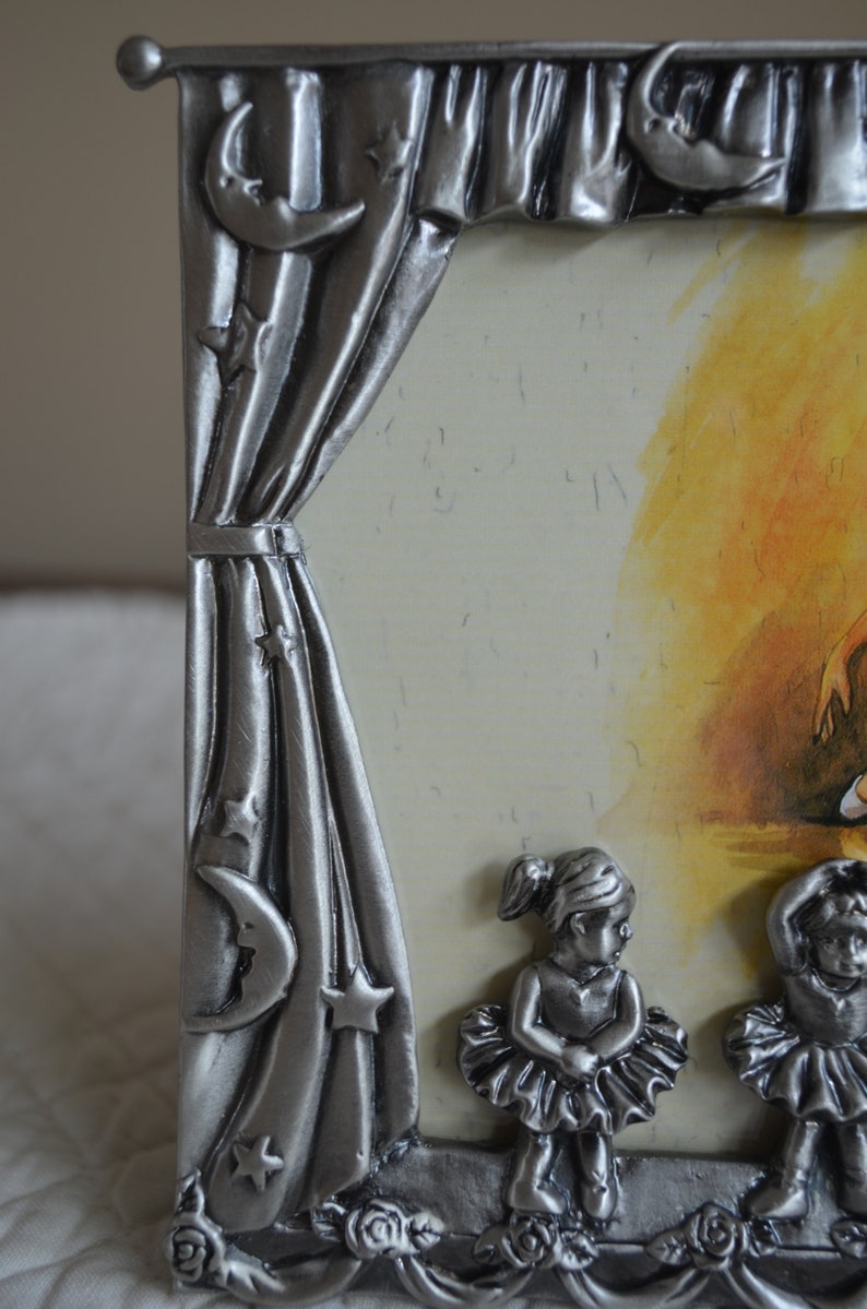 May include: Silver-tone picture frame with a curtain design and stars. The frame features three ballerina figurines in the bottom corners.