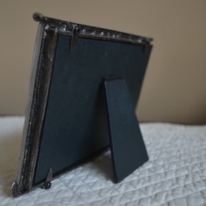 May include: A silver picture frame with a black backing and a fold-out stand. The frame has a decorative design around the edges.