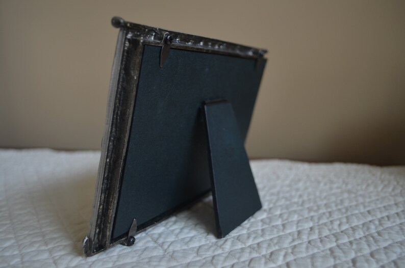 May include: A silver picture frame with a black backing and a fold-out stand. The frame has a decorative design around the edges.