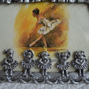 May include: A silver-tone picture frame with a ballerina image inside. The frame has a decorative design with a curtain and stars. Five small ballerina figurines are attached to the bottom of the frame.